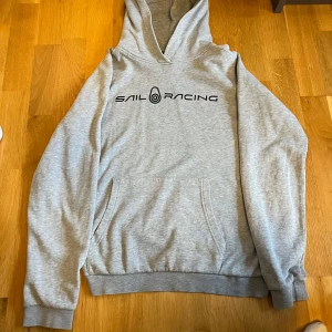 Grå hoodie från Sail Racing - Säljer en grå hoodie från Sail Racing med svart logotyp på bröstet. Tröjan har en klassisk design med känguruficka och ribbade muddar vid ärmslut och nederkant. Perfekt för en avslappnad stil.