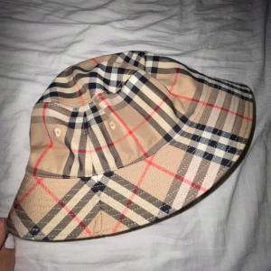 Burberry bucket hat - Snygg burberry bucket hat med klassiskt rutigt mönster i beige, svart och rött. Perfekt för att ge din outfit en stilren touch.