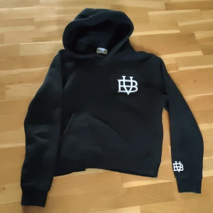 Svart hoodie från Bvaldi - Säljer en svart hoodie från Bvaldi med vit broderad logga på bröstet och ärmen. Den har en klassisk design med huva och långa ärmar. Perfekt för en avslappnad stil.