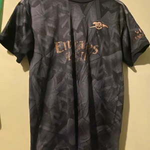 Svart fotbollströja med mönster - Snygg svart fotbollströja från Adidas med subtilt mönster och korta ärmar. Perfekt för fotbollsfans som vill ha en stilren look. Tröjan har en logotyp på bröstet och sponsortryck i guld.