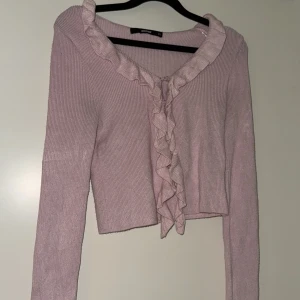 Ljusrosa stickad kofta med volangdetaljer - Säljer en söt ljusrosa stickad kofta med volangdetaljer längs halsringningen och framtill. Koftan har långa ärmar och en croppad passform, perfekt för en chic och feminin look. Passar bra till både jeans och kjol!