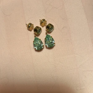Caroline Svedbom Örhängen - Eleganta guldörhängen med droppformade gröna stenar. Perfekta för att ge en touch av glamour till din outfit. Stiftfäste för enkel användning. Använda enstaka gånger då guld inte är min färg egentligen! Pris går att diskuteras. OBS! Har hund i hemmet men hon fäller INTE päls! Det i bakgrunden på bilden är från en bomullströja. 