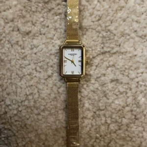 Guldfärgad klocka från Lorenzo Milano - Elegant guldfärgad klocka från Lorenzo Milano med fyrkantig urtavla och romerska siffror. Klockan har ett stilrent mesh-armband som ger en modern touch. Perfekt för att lyfta vilken outfit som helst.