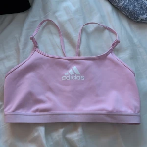Rosa sporttopp från Adidas - Säljer en rosa sporttopp från Adidas med smala axelband och korsad rygg. Toppen har en vit Adidas-logga framtill och elastiskt band med texten 'The Brand with the 3 Stripes'. Perfekt för träning eller vardagsbruk.