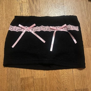 Svart kjol med rosa spetsdetaljer - Söt svart kortkjol med rosa spets och rosetter. Perfekt för en unik och lekfull stil. Kjolen har en tight passform och är gjord i ett mjukt material.