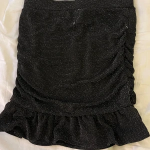 Svart glittrig kjol från Kappahl - Snygg svart glittrig kjol från Kappahl med volangdetalj i nederkant. Kjolen har en åtsittande passform och är perfekt för att ge en festlig touch till din outfit.
