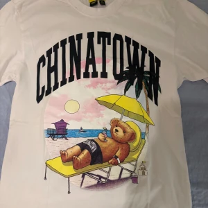 Vit t-shirt från Chinatown Market - Säljer en vit t-shirt från Chinatown Market med ett tryck av en nalle som solar på en strandstol under en palm. Texten 'CHINATOWN' är tryckt i stora bokstäver ovanför motivet. 