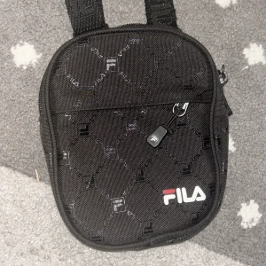 Svart axelväska från FILA - Snygg svart axelväska från FILA med ett diskret rutmönster och justerbar axelrem. Väskan har en praktisk dragkedja och är perfekt för att bära med sig det nödvändigaste. FILA-loggan syns tydligt på framsidan.