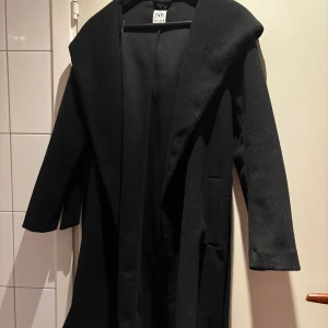 Svart kappa från Zara - Elegant svart kappa från Zara med långärmad design och knytband i midjan. Perfekt för höst och vinter, denna kappa ger en stilren look och är tillverkad i ett mjukt material.