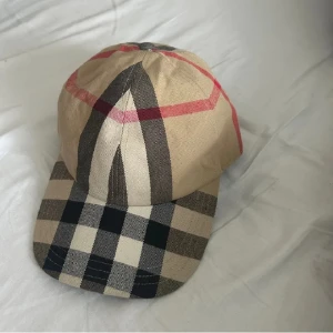 Beige rutig keps från Burberry - Snygg älta beige keps från Burberry med klassiskt rutigt mönster i svart och rött. Perfekt accessoar för en stilren look. Justerbar passform för optimal komfort.
