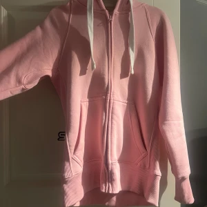 Russemerch Hoodie rosa  - Säljer en mysig rosa hoodie med dragkedja och stora fickor framtill. Den har långa ärmar och vita snören i huvan. Är knappt använd, varje fall inga tecken på användning 🩷nypris 749kr 