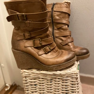Bruna kilklackade boots med spännen - Snygga bruna boots med kilklack och flera dekorativa spännen. Skorna har en dragkedja på sidan för enkel på- och avtagning samt en robust sula för bra grepp. Perfekta för höst och vinter. Fodrade