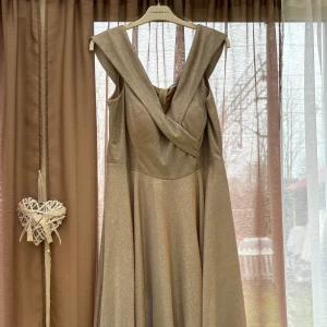 Champagnefärgad klänning med omlottdetalj - Elegant champagnefärgad klänning från Chi Chi London med omlottdetalj framtill. Klänningen har en glittrig yta och är ärmlös, vilket ger en sofistikerad look. Perfekt för speciella tillfällen.