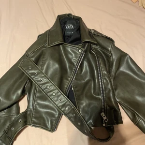 Olivgrön skinnjacka från Zara - Snygg olivgrön skinnjacka från Zara med dragkedja och bälte. Jackan har en cool bikerstil med långa ärmar och en klassisk krage. Perfekt för att ge din outfit en edgy touch, aldrig använt bara testat 1 gång och därför säljer jag den, pris kan diskuteras! 
