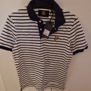 Oanvänd/ny Randig pikétröja från Lyle & Scott - Snygg randig pikétröja från Lyle & Scott i off-white och marinblått. Tröjan har korta ärmar och en klassisk krage med knappar. Perfekt för en stilren look.