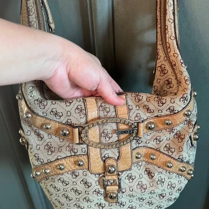 Guess  axelväska med monogram och nitar - Snygg beige axelväska från Guess med monogrammönster och dekorativa nitar. Väskan har detaljer i syntetmaterial som ger en lyxig känsla. Perfekt för att ge din outfit en extra touch. Väskan är endast använd ett fåtal gånger, därefter endast förvarats i garderoben. Finns ett par fläckar i innefoderet. 