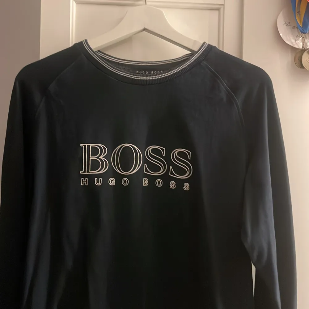 Snygg svart tröja från Hugo Boss med stort BOSS-tryck på framsidan. Tröjan har långa ärmar och en rund halsringning med diskreta ränder. Perfekt för en stilren look.. Neuletakit & Villapaidat.