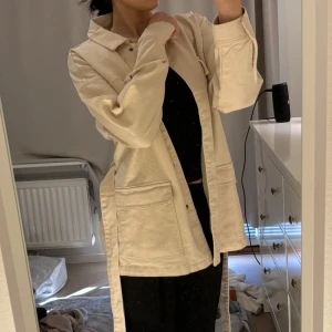 Beige overshirt kappa med knytband - Säljer en stilren beige overshirt med knappar och knytband i midjan. Jackan har långa ärmar och stora fickor framtill, perfekt för en avslappnad look.