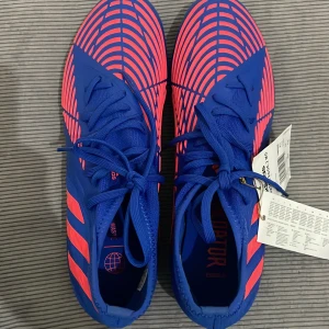Adidas Predator fotbollsskor - Snygga Adidas Predator fotbollsskor i blått med neonrosa detaljer. Skorna har en iögonfallande design med snörning och dobbar för optimalt grepp på planen. Perfekta för fotbollsspelare som vill sticka ut. Dem är helt nya och aldrig använt.