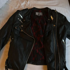 Svart skinnjacka från Jofama - Snygg svart skinnjacka från Jofama by Kenza med coola dragkedjedetaljer och en stilren design. Perfekt för att ge din outfit en edgy touch. Jackan har en asymmetrisk dragkedja och flera fickor med dragkedjor.