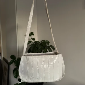 Vit axelväska med krokodilmönster - Säljer en superfin vit axelväska med krokodilmönster. Den har en stilren design och är perfekt för både vardag och fest. Väskan är i syntetmaterial och har en praktisk storlek för att få plats med det viktigaste.