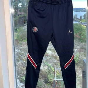 Svarta träningsbyxor från Jordan - Säljer ett par svarta träningsbyxor från Jordan med PSG-logga. Byxorna har en snygg design med röda och vita ränder vid benen och en dragkedja vid vristen för justerbar passform. Perfekta för träning eller en avslappnad stil.