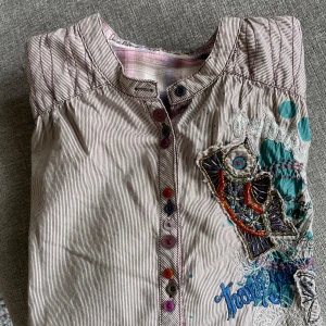 Desigual blus i nyskick - Snygg skjorta med långa ärmar och en unik design. Den har en mörkbrun och vit randig bakgrund med en stor broderad detalj på framsidan. Skjortan har en knappslå och en rundad krage. Den är i nyskick. •Storlek L Djurfritt och rökfritt hem 