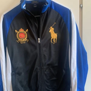 Svart och blå jacka från Polo Ralph Lauren - Säljer en snygg svart och blå jacka från Polo Ralph Lauren. Jackan har en dragkedja framtill och är dekorerad med broderade emblem i guld och rött på bröstet. Den har långa ärmar och är perfekt för en sportig look. Passar både till vardags och till mer avslappnade tillfällen.