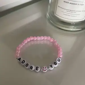 Säljer ett supergulligt armband med rosa pärlor och vita bokstavspärlor som bildar texten 'ADORE YOU'. Det finns också en söt smiley på en av pärlorna. Perfekt för att ge en personlig touch till din outfit!