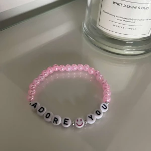 Rosa pärlarmband med text - Säljer ett supergulligt armband med rosa pärlor och vita bokstavspärlor som bildar texten 'ADORE YOU'. Det finns också en söt smiley på en av pärlorna. Perfekt för att ge en personlig touch till din outfit!