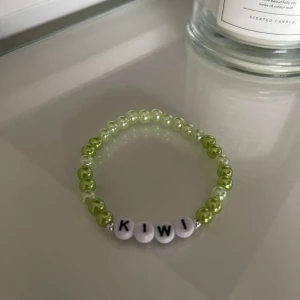 Pärlarmband med texten 'KIWI' - Snyggt och färgglatt armband med pärlor i gröna och vita nyanser. Armbandet har bokstavspärlor som bildar ordet 'KIWI'. Perfekt accessoar för att liva upp vilken outfit som helst!