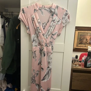 Rosa blommig klänning - Säljer en superfin rosa klänning med blommigt mönster. Den har en omlott-design med korta ärmar och ett knytband i midjan. Perfekt för sommarens fester eller en dag på stan!