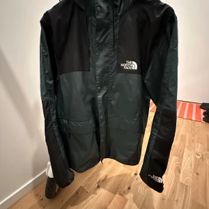 Svart och grön jacka från The North Face - Säljer en snygg svart och grön jacka från The North Face.Den är perfekt för utomhusaktiviteter och har en praktisk huva. Jackan är tillverkad i 100% polyester och har en stilren design med The North Face-loggan på bröstet och ryggen. Den är i bra s