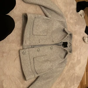 Grå kavaj från 157 - Säljer en snygg grå kapp från märket  i storlek XS. Den är i nyskick och har en stilren design med knappar framtill. Perfekt för höst och vår, och funkar både till vardags och fest. Kappan har långa ärmar och är gjord i ett mjukt material som kän