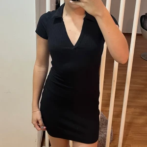 Svart kortklänning med krage - Säljer en stilren svart kortklänning med krage och v-ringning. Klänningen har en tight passform och korta ärmar, perfekt för en utekväll eller fest. Materialet är mjukt och stretchigt, vilket gör den bekväm att bära hela dagen.