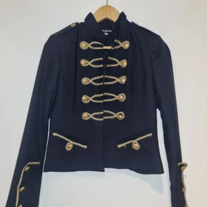 Svart kavaj med gulddetaljer - Säljer en snygg svart kavaj från Rebel Cashmere med coola gulddetaljer. Den har långa ärmar och är dekorerad med guldknappar och broderade detaljer fram och på ärmarna. Perfekt för en festlig kväll eller när du vill sticka ut lite extra. Kavajen är i mycket bra skick och har en elegant passform. Stolek 36/S. Oanvänd utan etikett. 