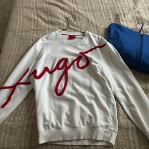 Vit tröja från Hugo - Säljer en snygg vit tröja från Hugo med röd text på framsidan. Tröjan har en klassisk rund hals och är perfekt för både vardag och fest. Den är i bra skick och redo för en ny ägare. Passar perfekt till jeans  för en stilren look!