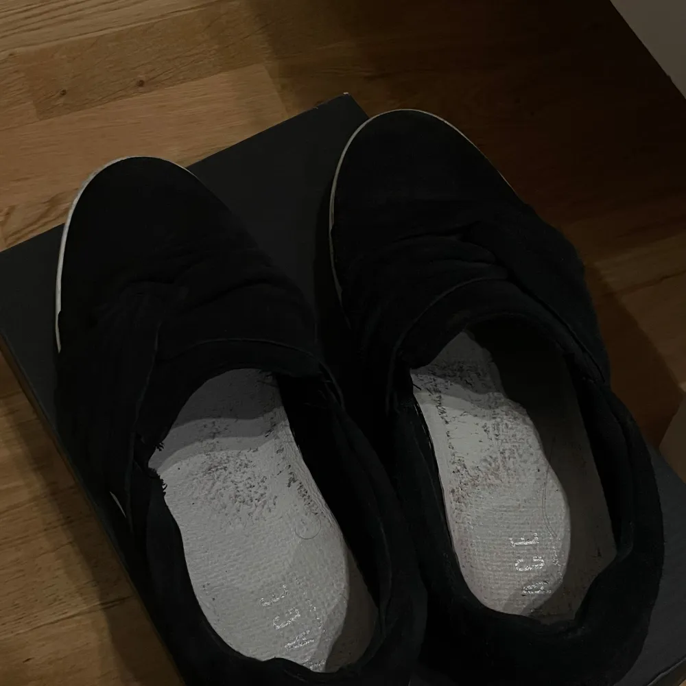 Säljer ett par svarta sneakers i mocka med vit sula. De har en stilren design utan snörning, vilket gör dem enkla att ta på och av.. Skor.