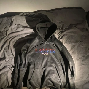 Svart hoodie från Lanvin - Säljer en snygg svart hoodie från Lanvin Paris. Den har en klassisk design med en stor ficka fram och en bekväm huva. Perfekt för både vardag och chill. Texten på bröstet är i rött och blått, vilket ger en cool kontrast. Passar perfekt till jeans eller joggers!