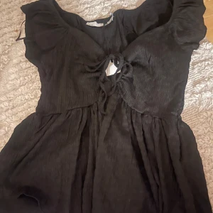 Svart topp med knytning - Säljer en svart topp i storlek XS. Den har en snygg knytning framtill och är gjord i ett mjukt, lätt material. Perfekt för både vardag och festliga tillfällen. Toppen har korta ärmar och en bekväm passform. Passar bra till jeans eller kjol för en stilren look.