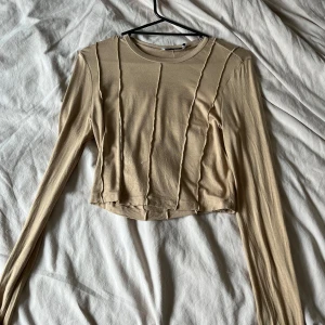 NA-KD beige tröja  - En beige långärmad t-shirt med sömmar på framsidan. T-shirten är cropped och har en rund halsringning. Väldigt stretchiga material, super skön! 