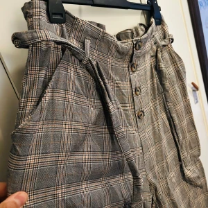 Rutig byxa från H&M - Säljer en superfin rutig byxa från H&M i storlek XL. Byxorna är högmidjade med knappar framtill och ett matchande bälte. Perfekt för höst och vår med sitt klassiska rutmönster i grått och beige. Tillverkad i 100% bomull vilket gör dem både bekväma och stiliga. Passar perfekt för en chic vardagslook!