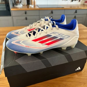 Adidas F50 league - Adidas F50 League FG/MG fotbollsskor i vit, blå och röd färg. Skorna är i mycket bra skick (se bilder) och har endast använts på två träningar men är tyvärr en storlek för stora för sonen. De är perfekta för både gräs och konstgräs. Strl 43 & 1/3