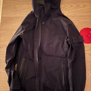 Svart jacka från Stone Island - Säljer en stilren svart jacka från Stone Island med huva och dragkedja. Jackan har en praktisk ficka på bröstet och en ikonisk logga på ärmen. Perfekt för höst och vår.