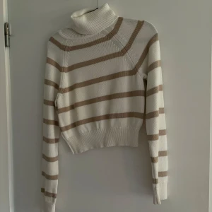 Randig polotröja i beige och vit - Säljer en mysig randig polotröja i beige och vit. Tröjan har långa ärmar och en hög polokrage, perfekt för kyligare dagar. Den är croppad och har ribbade muddar vid ärmslut och nederkant.