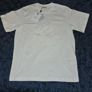 Vit t-shirt från Moncler - Snygg vit t-shirt från Moncler med ett unikt upphöjt mönster på framsidan. T-shirten har korta ärmar och ett broderat Moncler-märke på ärmen. Perfekt för en stilren look.