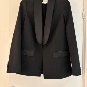 Svart kavaj från H&M - Elegant svart kavaj från H&M i ullblandning. Kavajen har en klassisk design med knappar och fickor framtill. Perfekt för en stilren look.