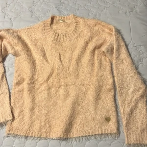 Rosa fluffig tröja - Mysig och fluffig rosa tröja med rund halsringning. Perfekt för kyliga dagar när du vill ha något bekvämt och stilrent. Tröjan har långa ärmar och en liten hjärtdetalj nedtill. Passar bra till både jeans och kjol.