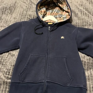 Mörkblå huvtröja Ralph lauren  - Säljer en mörkblå huvtröja från ralph Lauren med dragkedja och ett klassiskt rutigt foder i huvan. Tröjan har en liten logga på bröstet och praktiska fickor framtill. Perfekt för en avslappnad stil.