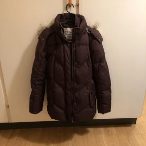 Brun dunjacka från Esprit - Säljer en brun dunjacka från Esprit med pälskantad huva. Jackan har dragkedja och långa ärmar, perfekt för kalla vinterdagar. Den är både stilren och funktionell.